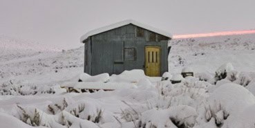 A small cabin made of corrugated steel and a yellow front door sits in sagebrush hills covered with snow and a sunset.