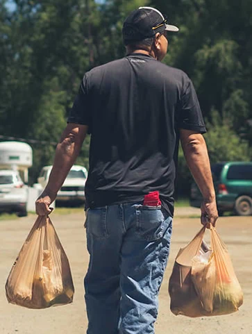 Man walking away from the camera holding two grocery bags full of food in a parking lot.