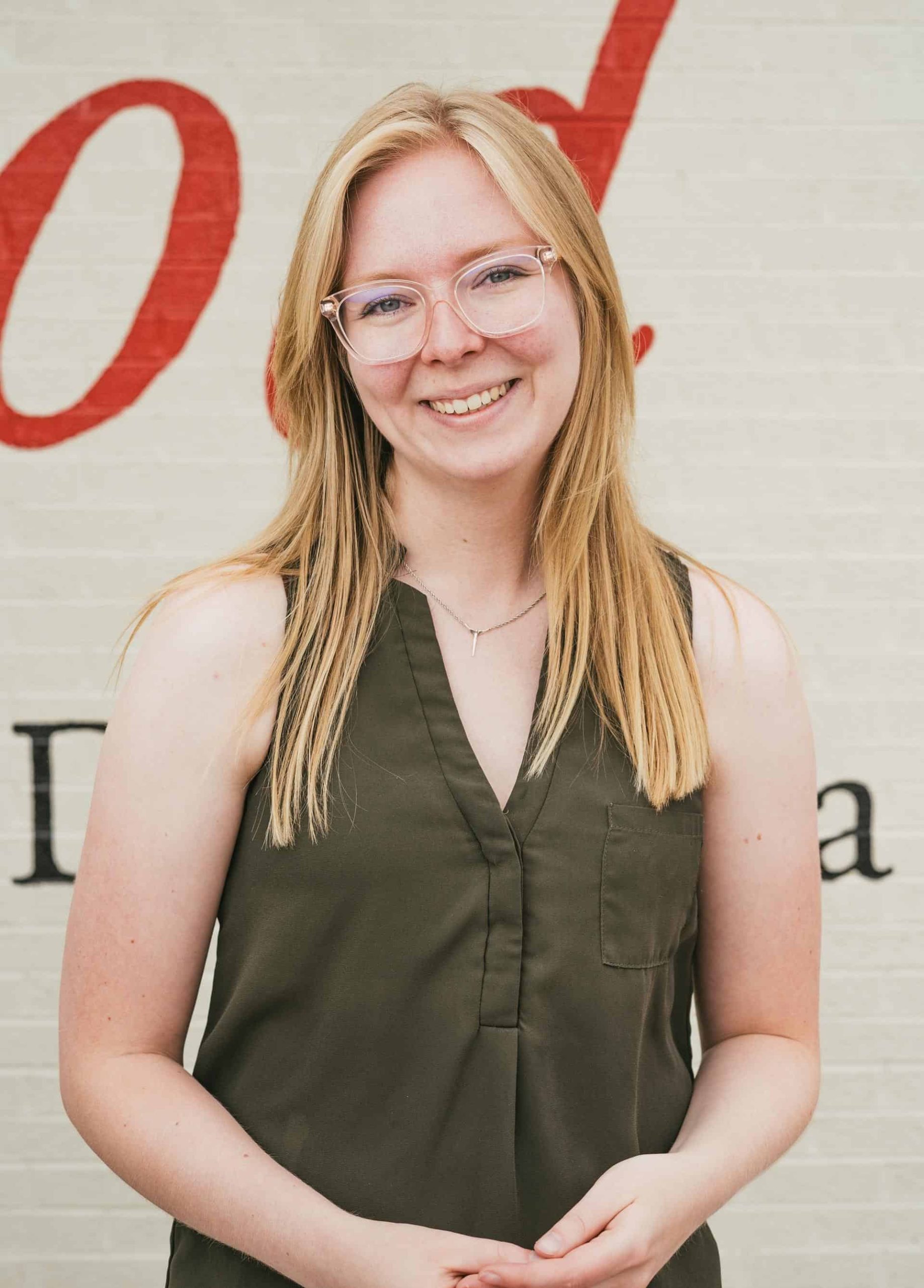 McKenzie Mathewson stands in front of the Gunnison Country Food Pantry wearing an olive blouse, glasses, and long blond hair.